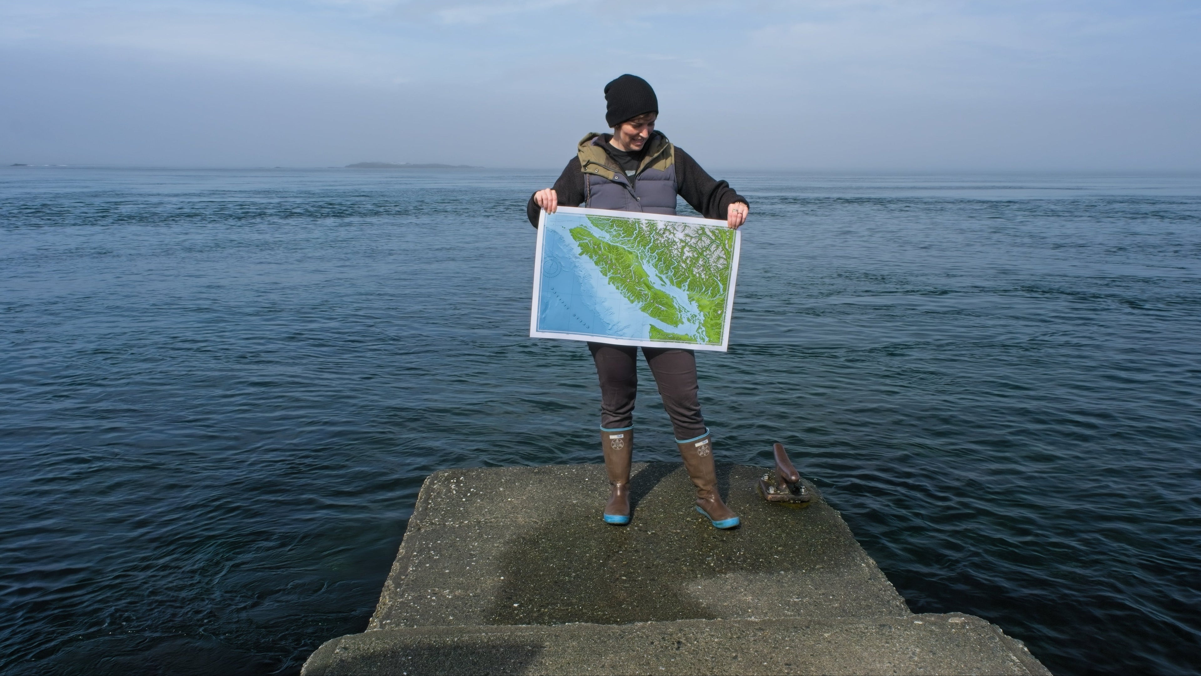 Person standing on a jetty, holding a map of Vancouver Island, surrounded by blue water water and a hazy sky