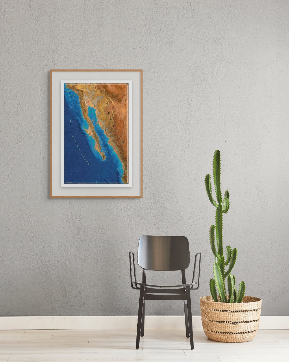 Framed artwork of a map of Baja and the Sea of Cortes on a wall with a chair and potted cactus below.