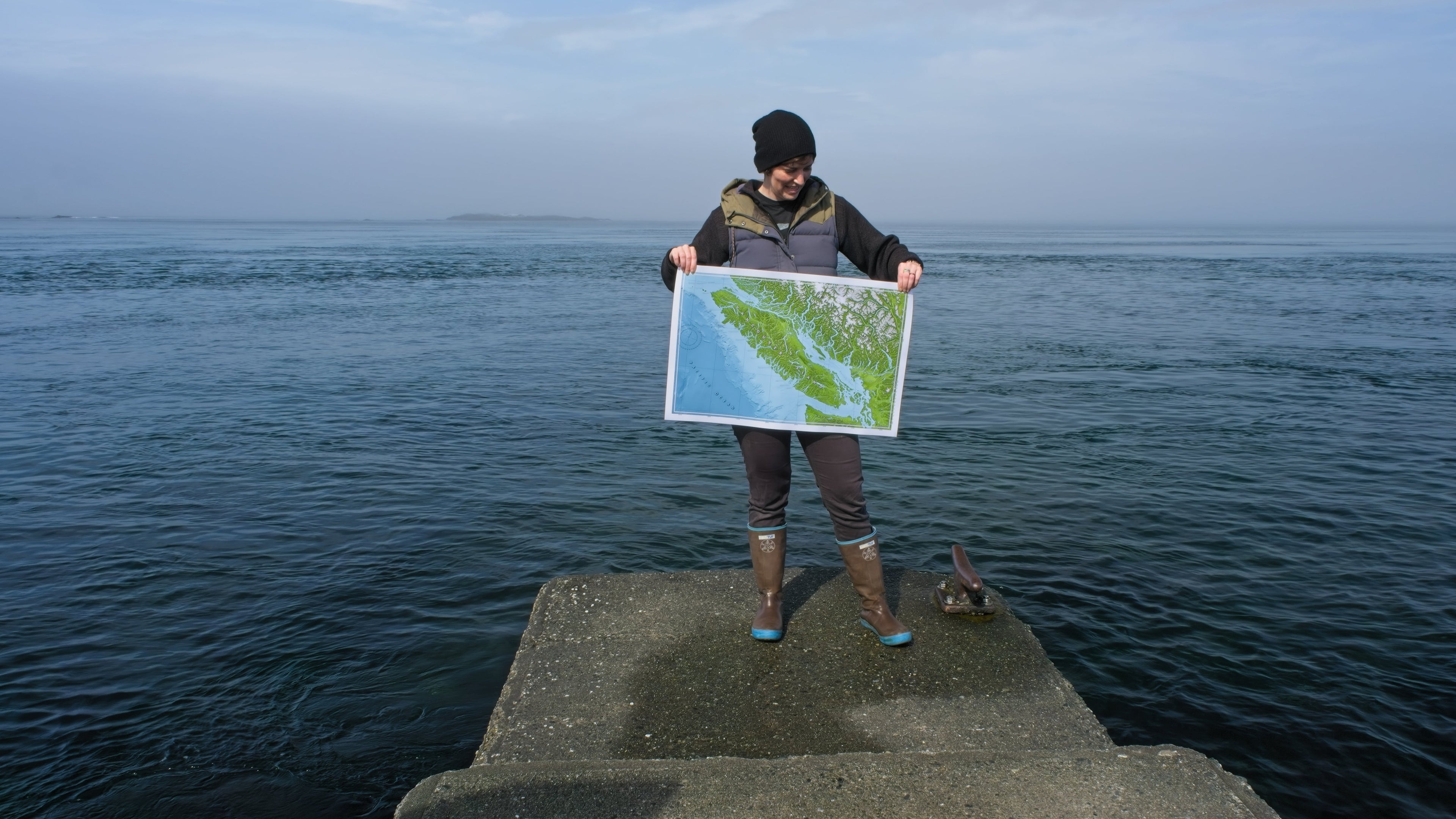 Person standing on a jetty, holding a map of Vancouver Island, surrounded by blue water water and a hazy sky