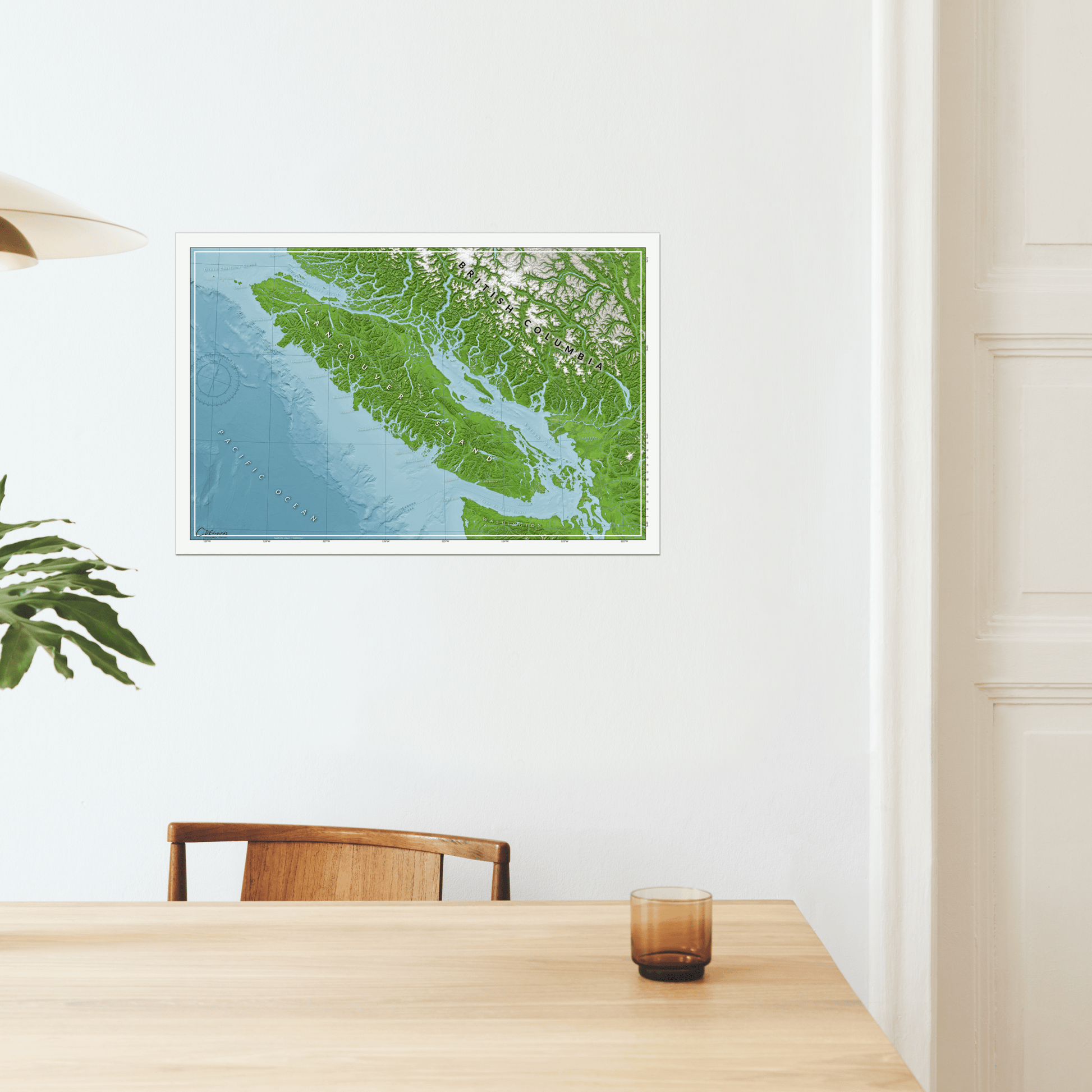 A printed map poster for wall art of Vancouver Island displayed, with various shades of green and blue representing land and water features. Hanging in a minimalist dinning room.