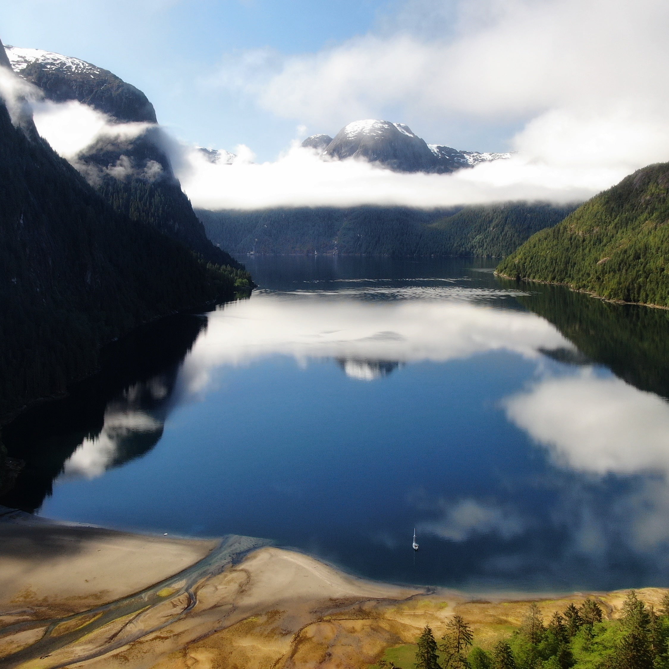 Deep ocean fjord surrounded by mountains with clouds reflecting on the water and a small sailboat anchored close to shore. 
