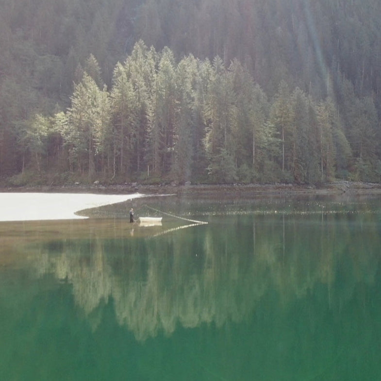 Person standing next to a white dinghy at the edge of the ocean in the Pacific Northwest, flyfishing out into an emerald green sea