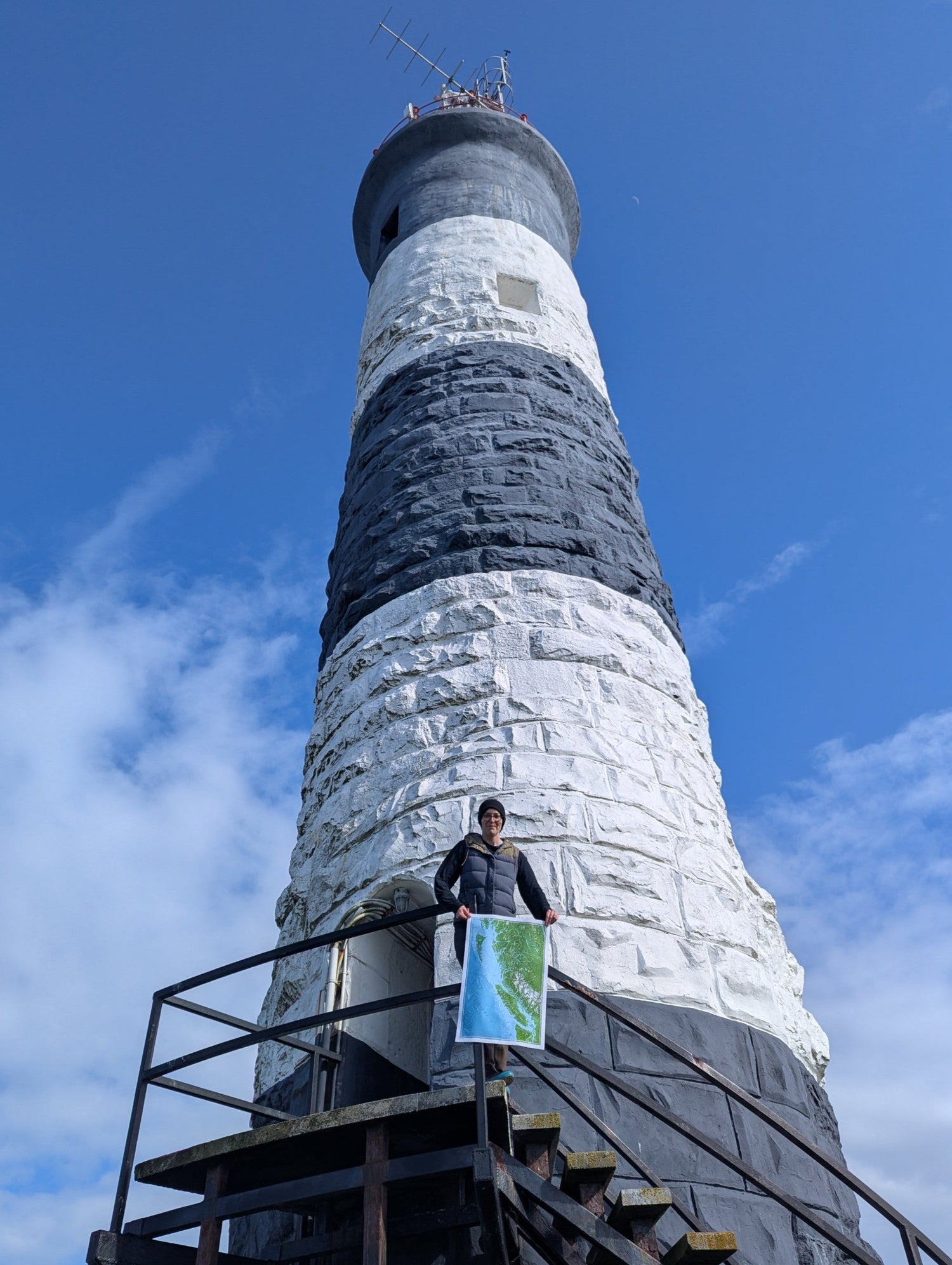 Person holding a map of British Columbia, standing on a platform at the base the Race Rocks lighthouse against a blue sky with clouds.