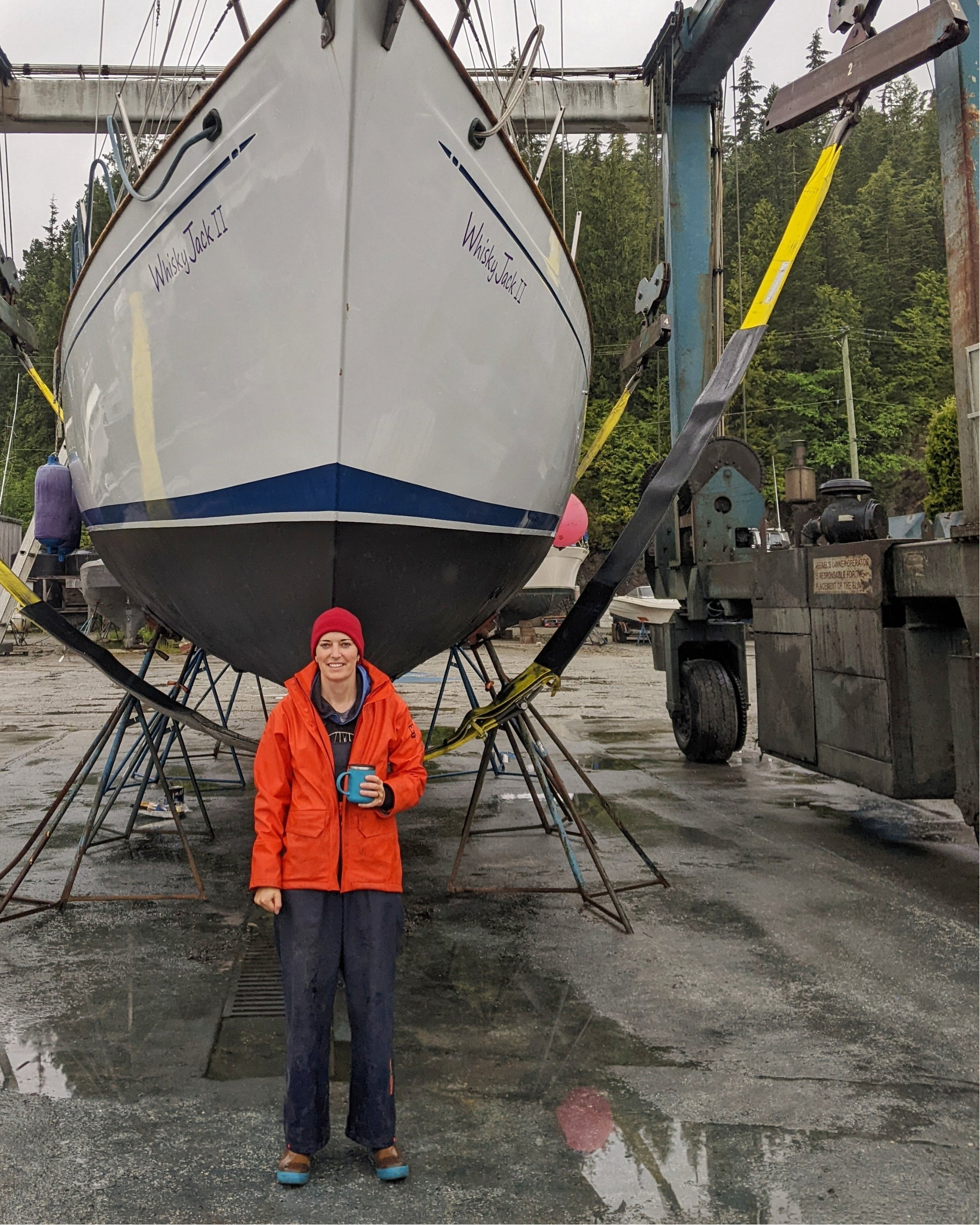 A white hulled sailboat hauled out on land with a person in an orange raincoat standing in front of it