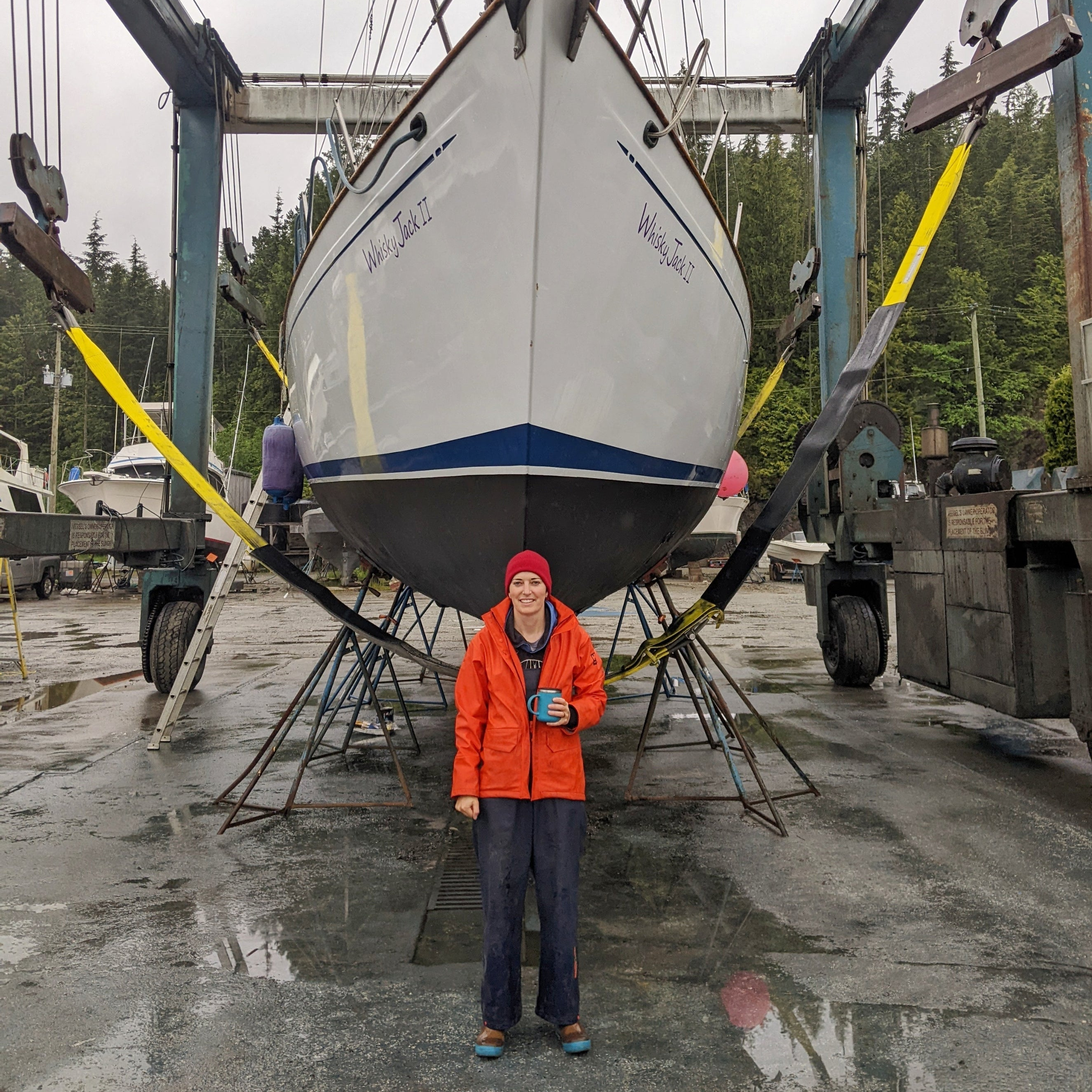 A white hulled sailboat hauled out on land with a person in an orange raincoat standing in front of it