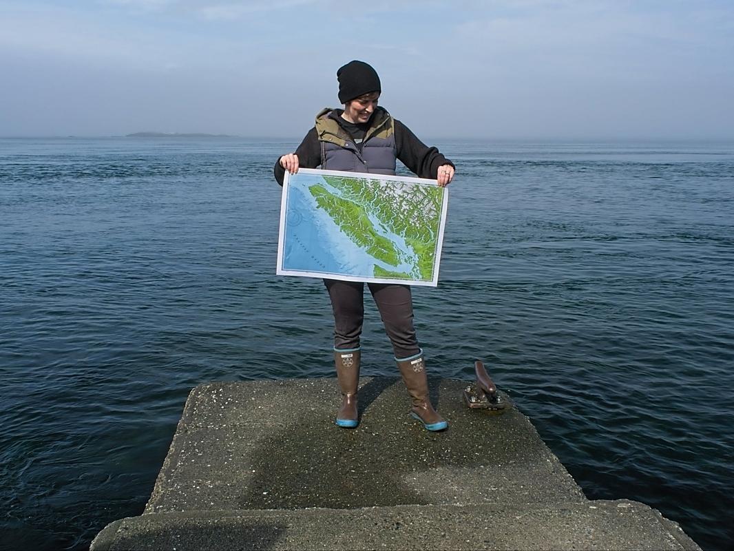 Person wearing gumboots standing on the edge of a concrete jetty holding a sailors map of Vancouver Island.